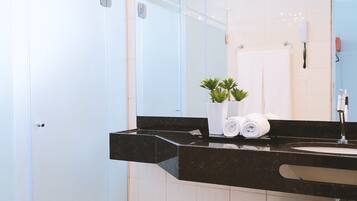 Un baño moderno con cabina de ducha de vidrio, encimera negra, banco de madera y suelo de baldosas blancas.