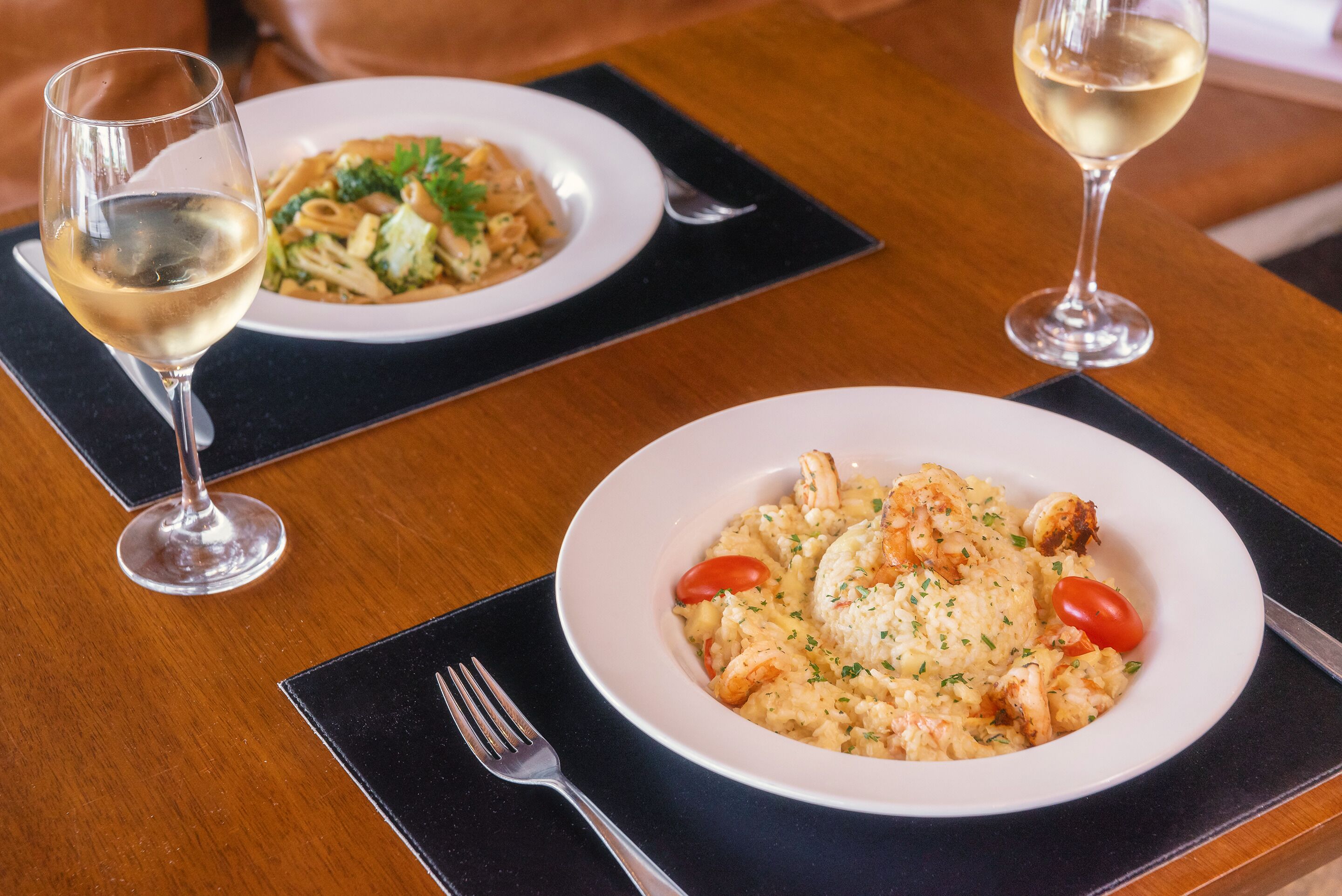 Dos platos de pasta y dos copas de vino blanco sobre una mesa de madera.