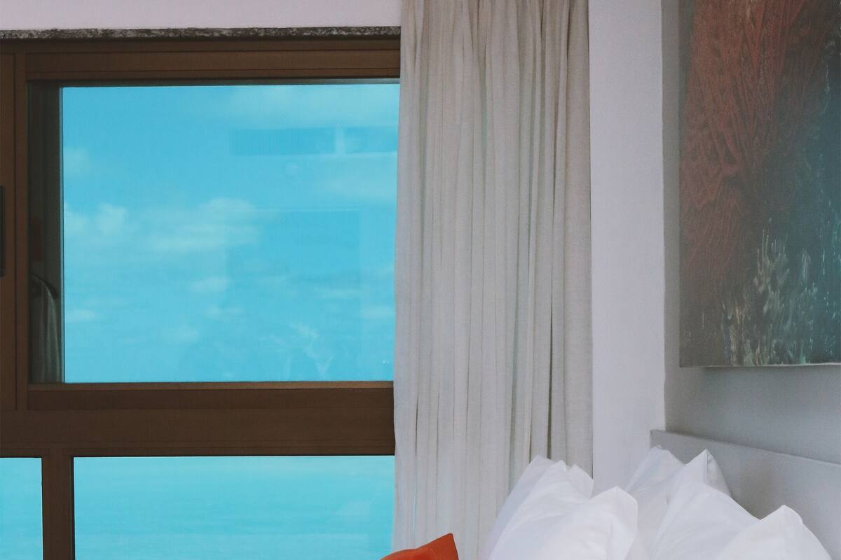 A bedroom with a large window, white bedding, and a view of the ocean.