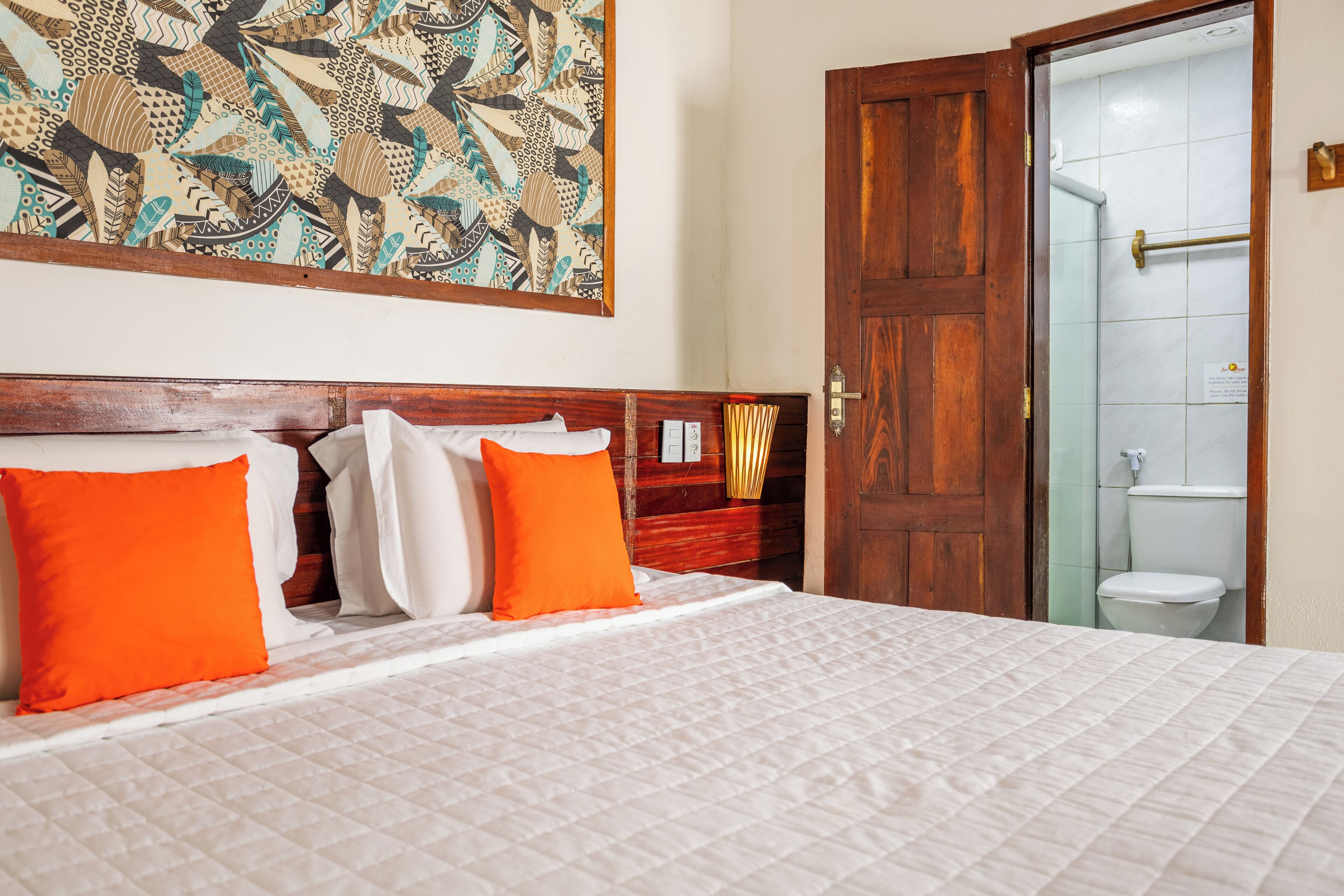 Habitación de hotel con una cama grande, dos almohadas naranjas, un cabecero de madera y un tapiz con motivos decorativos.