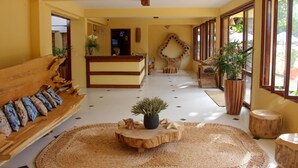 A hotel lobby with a reception desk, a wooden bench with cushions, a large woven rug, and potted plants. - Jeri Village Hotel (Jijoca de Jericoacoara)