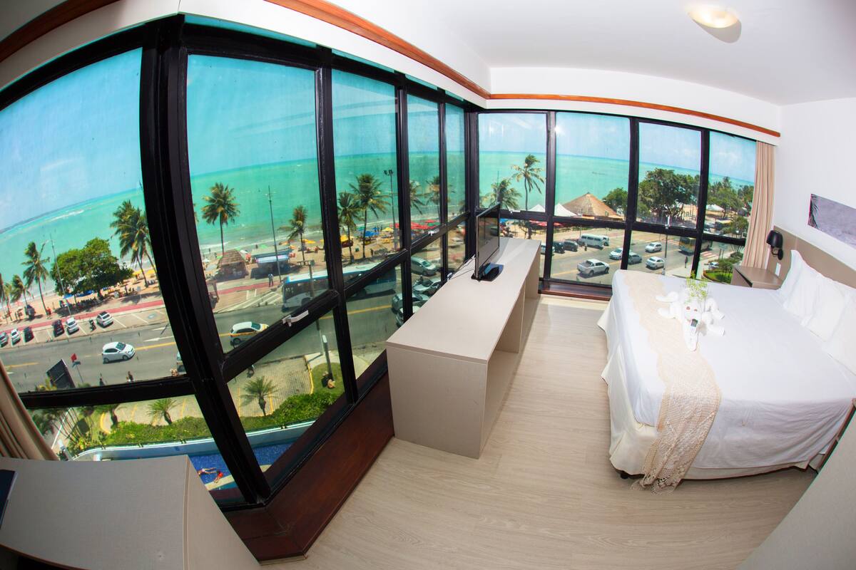 Une chambre d’hôtel dotée d’une grande baie vitrée panoramique, d’un lit, d’une petite table et offrant une vue sur la plage et l’océan.