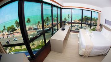 Quarto de hotel com uma grande janela panorâmica, uma cama, uma mesinha e vista para a praia e o oceano.