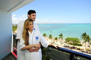 Ein Paar steht auf einem Balkon mit Blick auf einen Strand, Palmen und einen klaren Himmel.