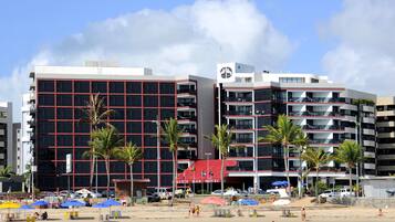 Un hotel con varios pisos, balcones y una zona de playa con sombrillas y palmeras.