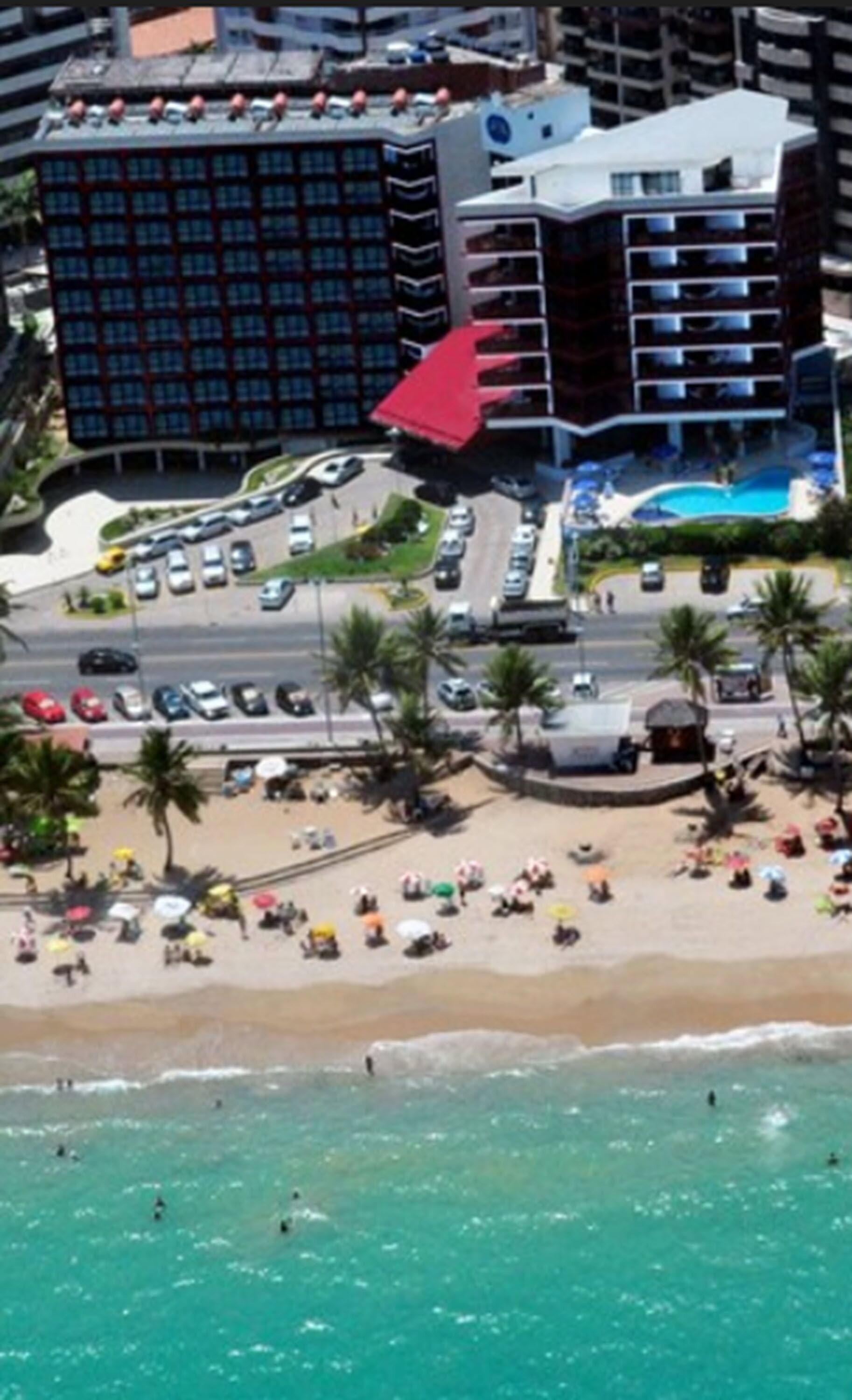 A coastal hotel with multiple floors, a parking area, and a beach with umbrellas.