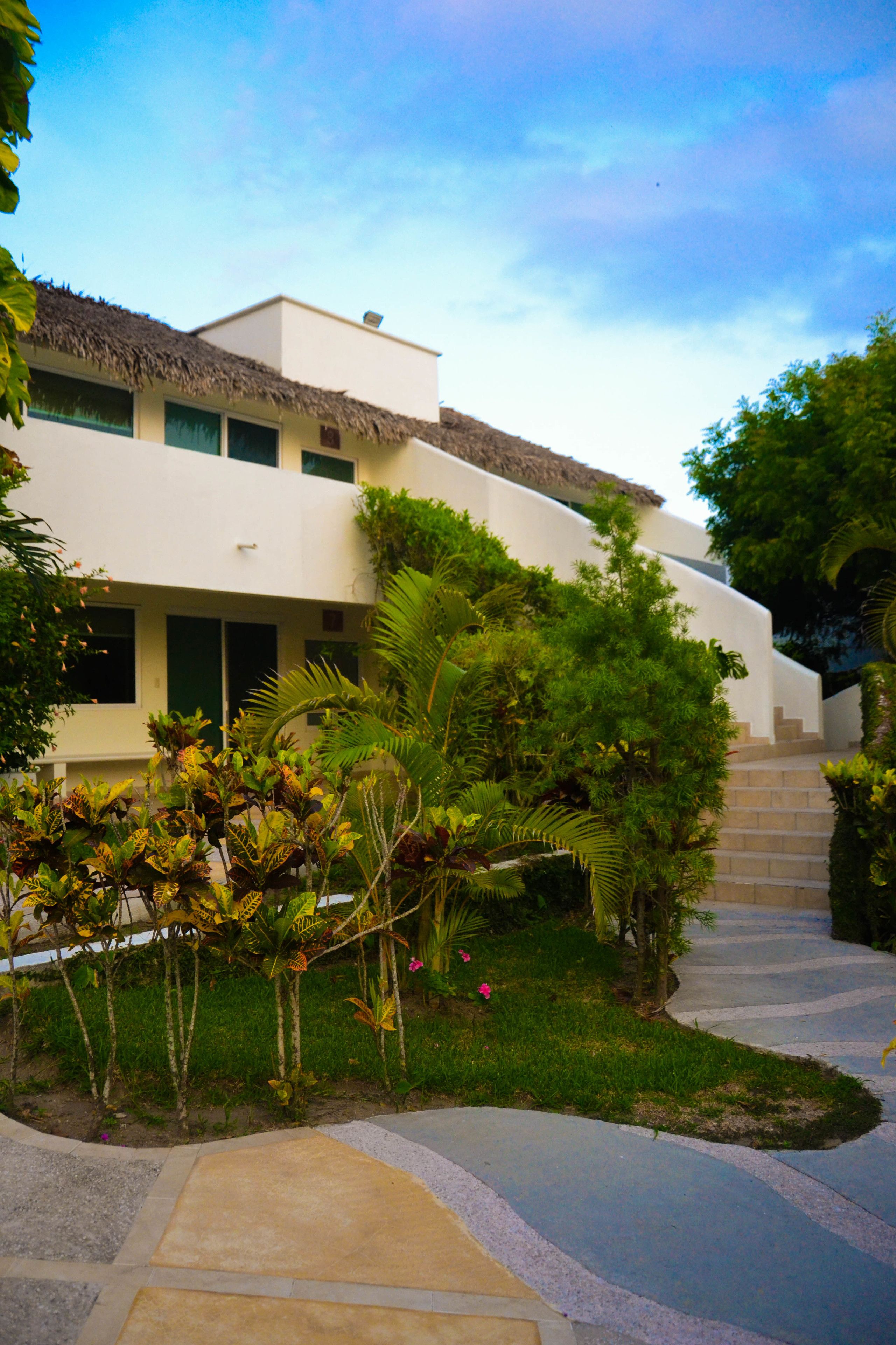 A modern building with a flat roof and a thatched roof section, surrounded by lush greenery and a stone pathway leading to the entrance.