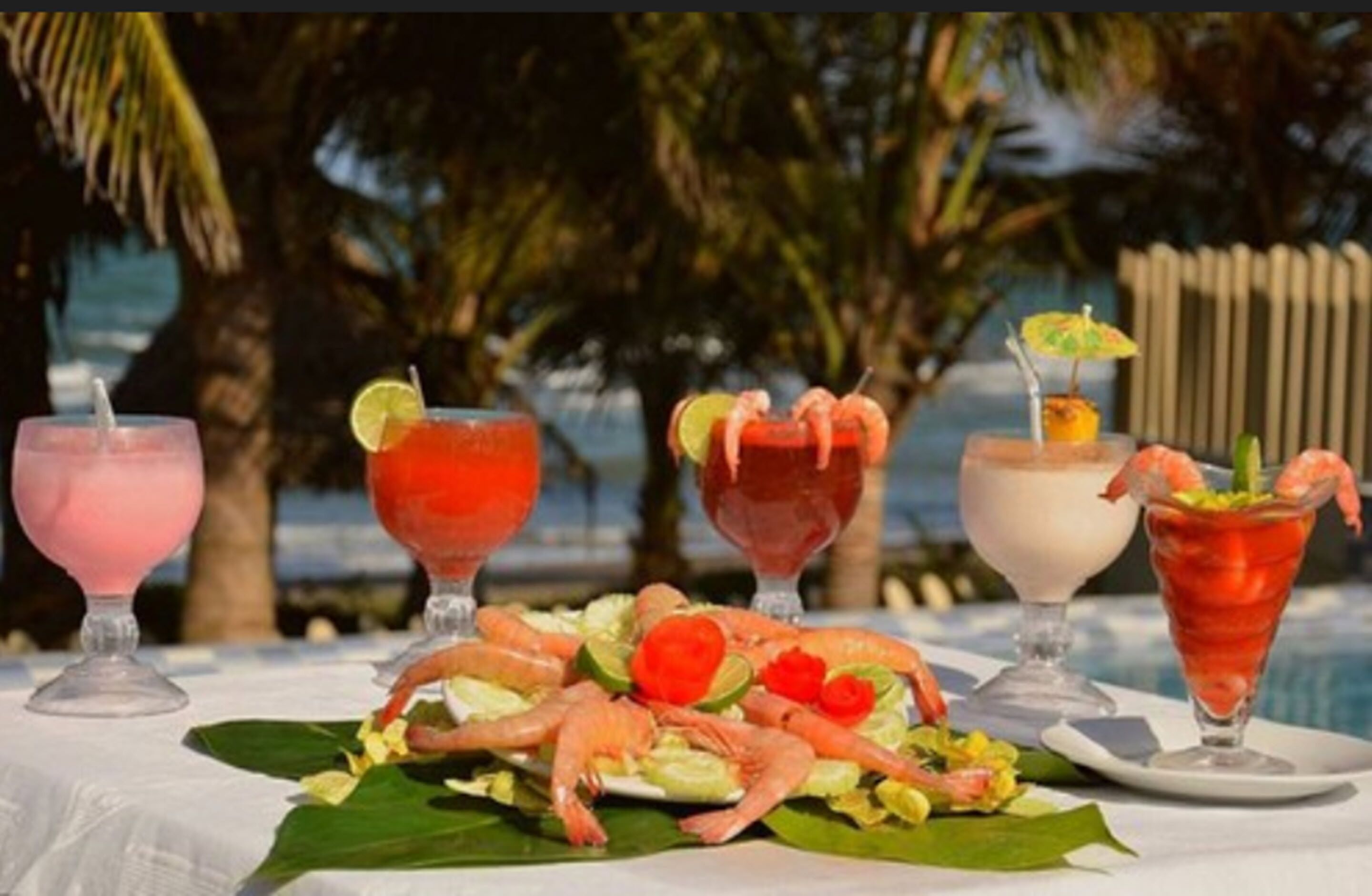A table with four drinks and a plate of shrimp and salad, set against a tropical background.