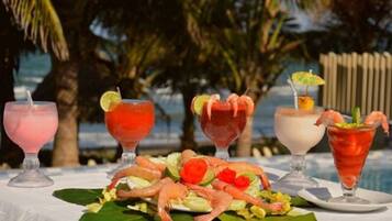 A table with four drinks and a plate of shrimp and salad, set against a tropical background.