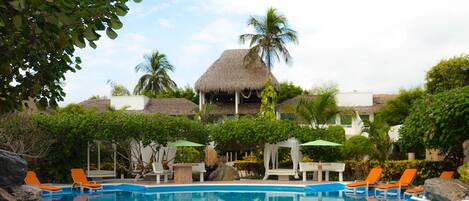 A swimming pool with lounge chairs, a thatched-roof structure, and tropical vegetation.