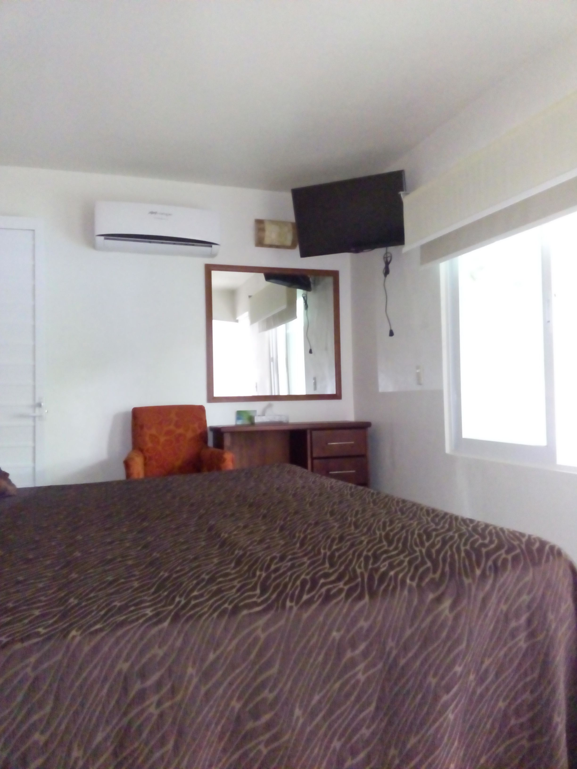 A hotel room with a large bed, a chair, a dresser, an air conditioning unit, a television mounted on the wall, and a window providing natural light.