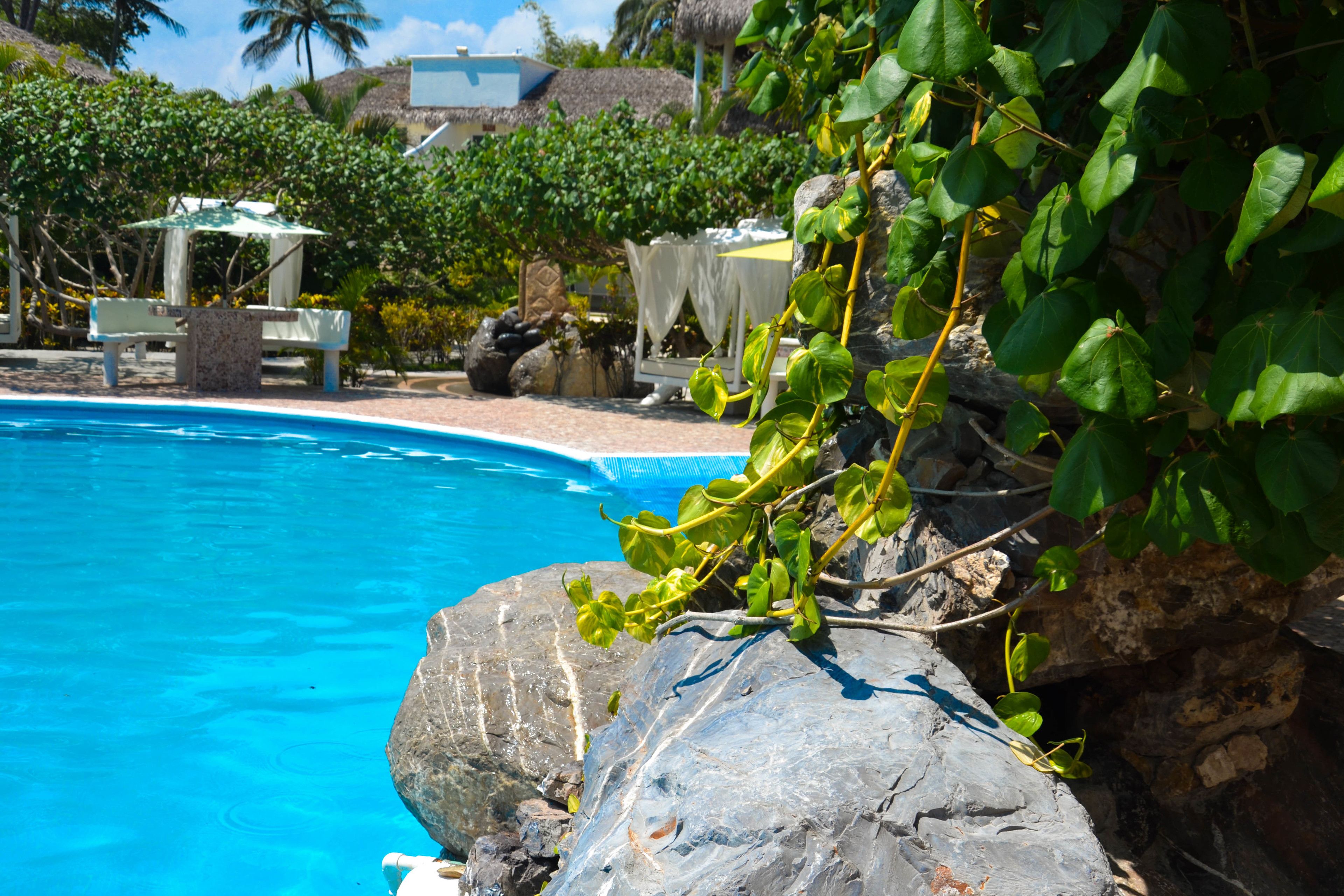 A clear blue pool surrounded by rocks and greenery, with a thatched-roof structure and a white table in the background.
