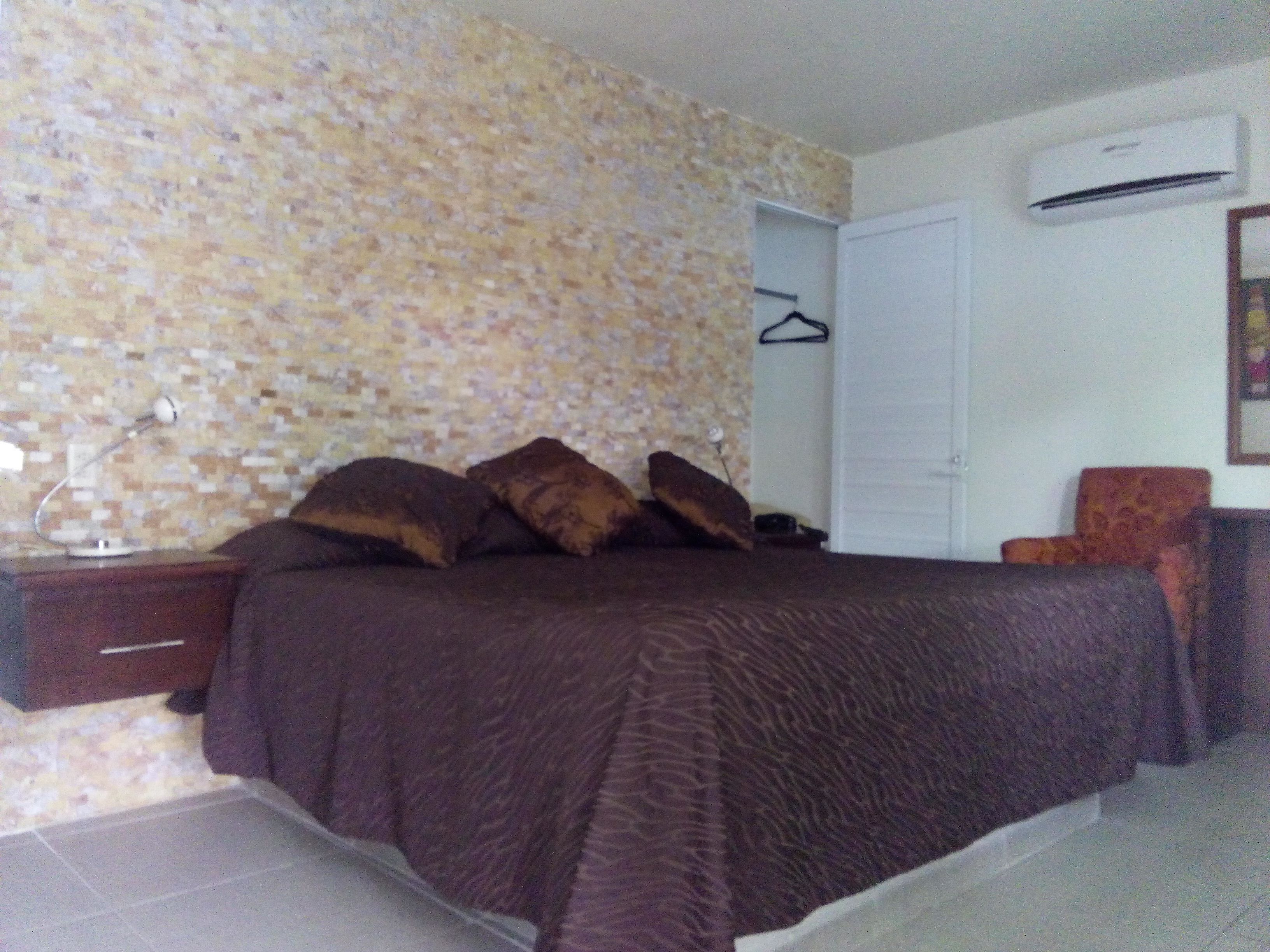 Un dormitorio con pared estampada, una cama con cubrecama marrón, una mesita de noche, una silla y un equipo de aire acondicionado.