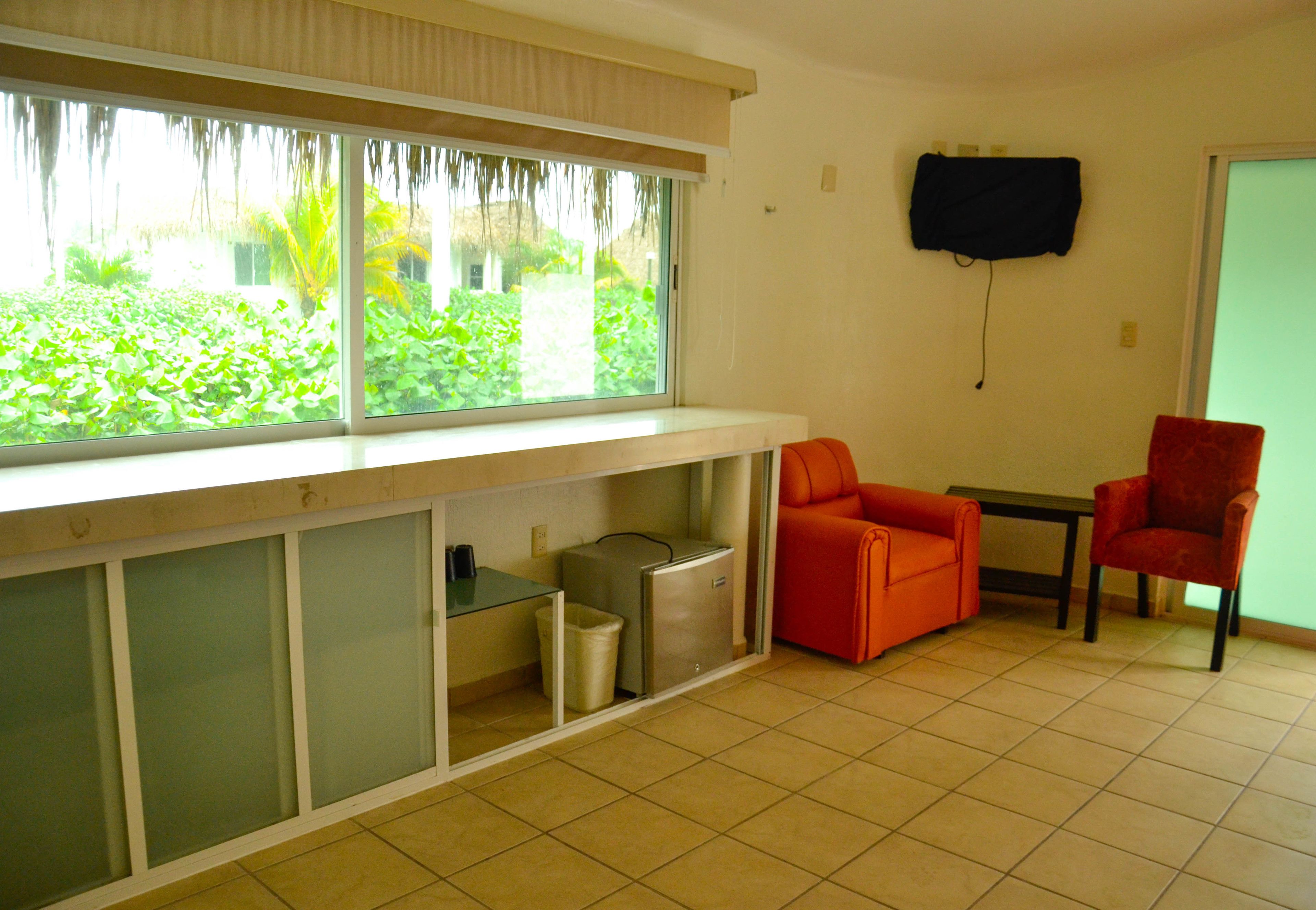 A room with a window, a television mounted on the wall, a trash bin, and two chairs.