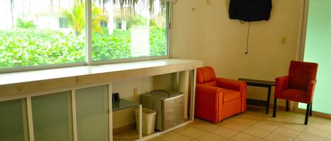 A room with a window, a television mounted on the wall, a trash bin, and two chairs.