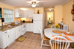 Una cocina con armarios blancos, una estufa, un microondas y un refrigerador. Hay una mesa de comedor con sillas y un florero con flores.