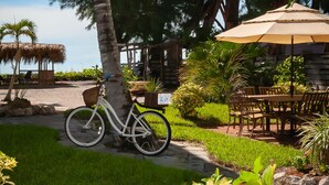 A bicycle parked by a palm tree, outdoor seating area with tables and chairs, and a thatched-roof structure.