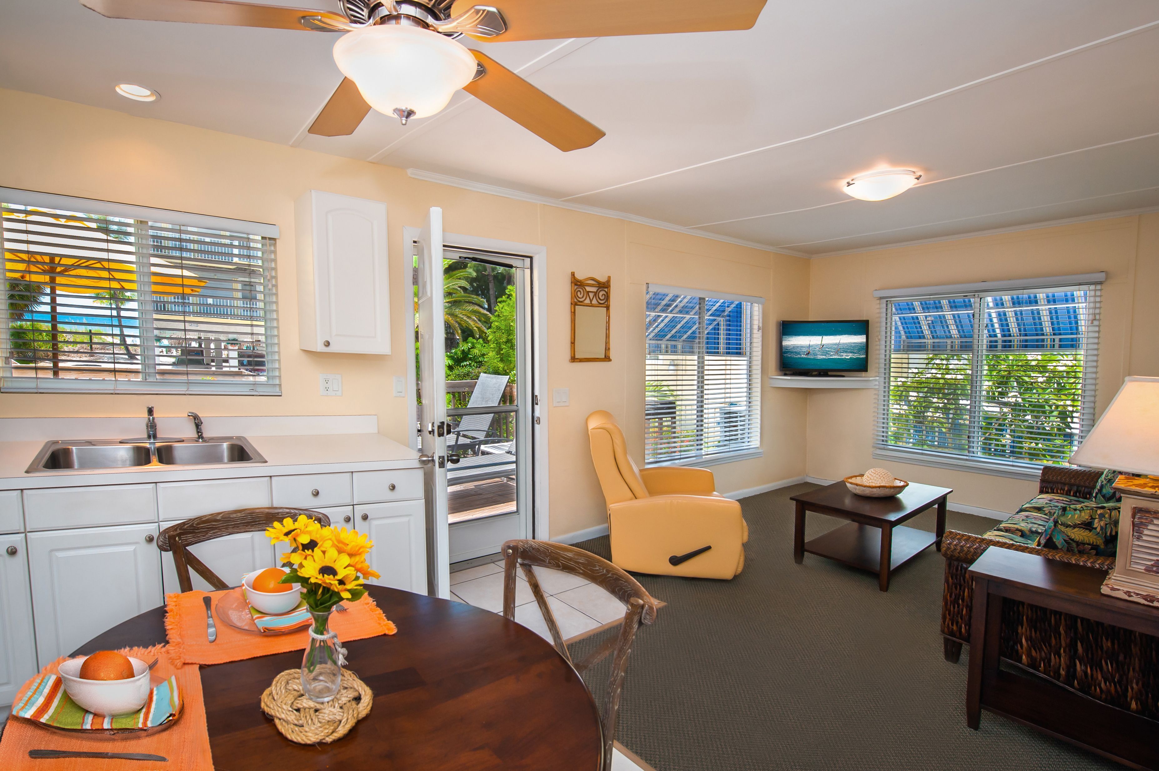 A modern living room with a dining area, a ceiling fan, a television, and a view of the outdoors through large windows.