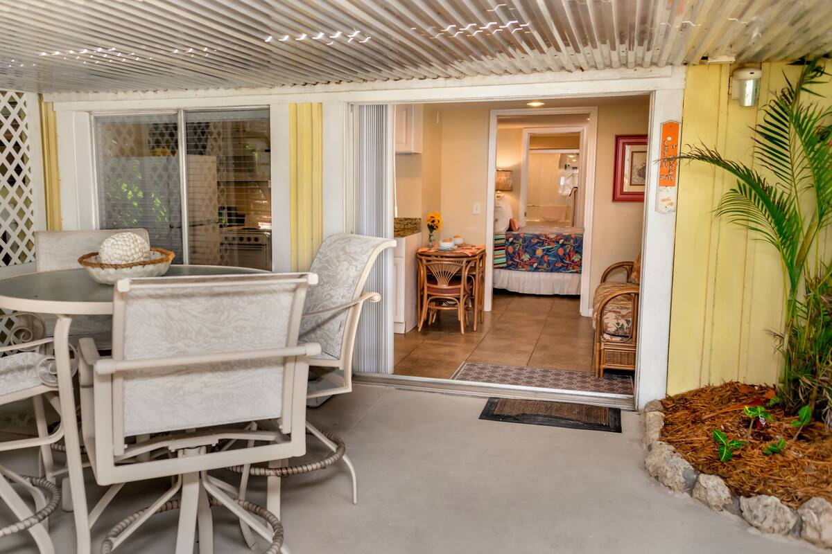 Een eetgedeelte met een ronde tafel en stoelen, een bijzettafeltje met een vaas, en uitzicht op een slaapkamer met een bed en een stoel.