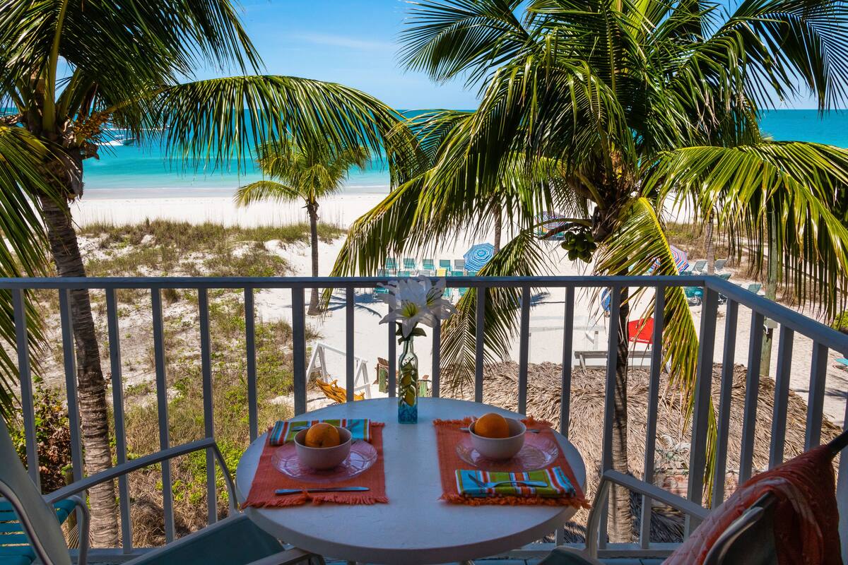 Een balkon met een tafel gedekt voor twee, met uitzicht op een strand met palmbomen en een helderblauwe oceaan.