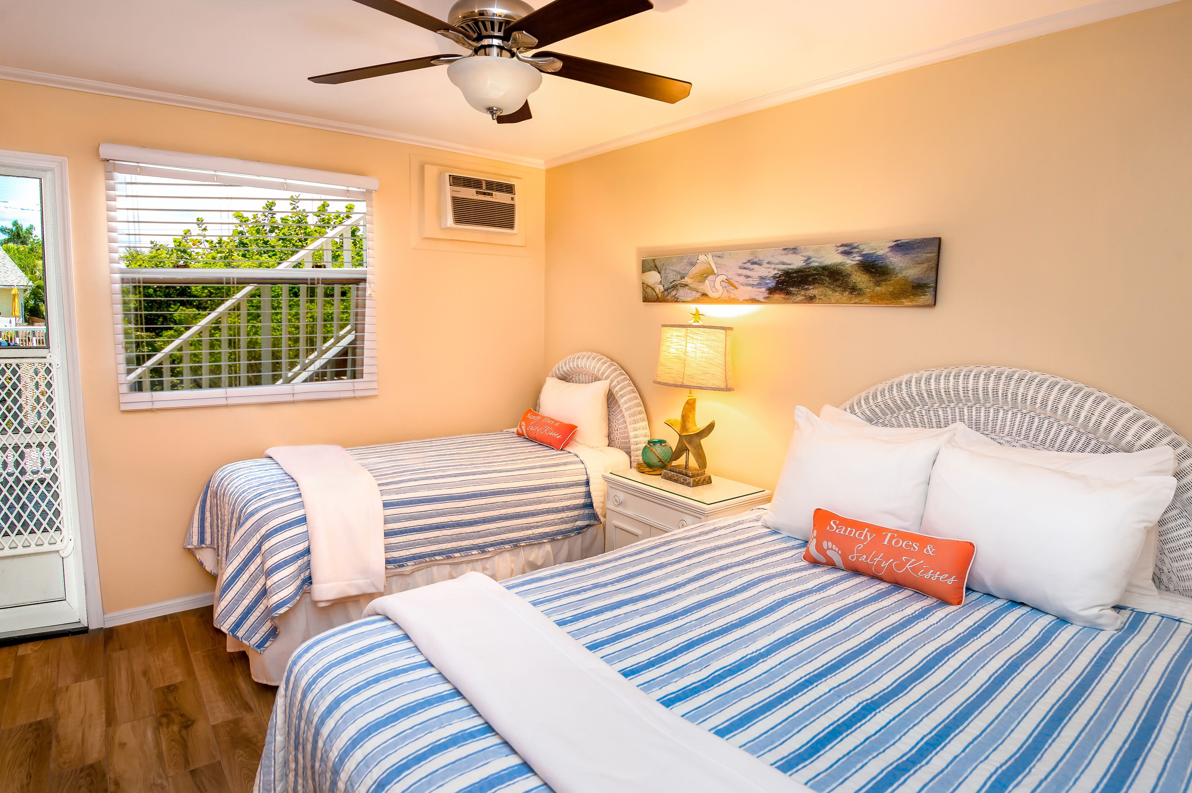 A bedroom with two beds, a ceiling fan, a window with blinds, a nightstand with a lamp, and a painting on the wall.