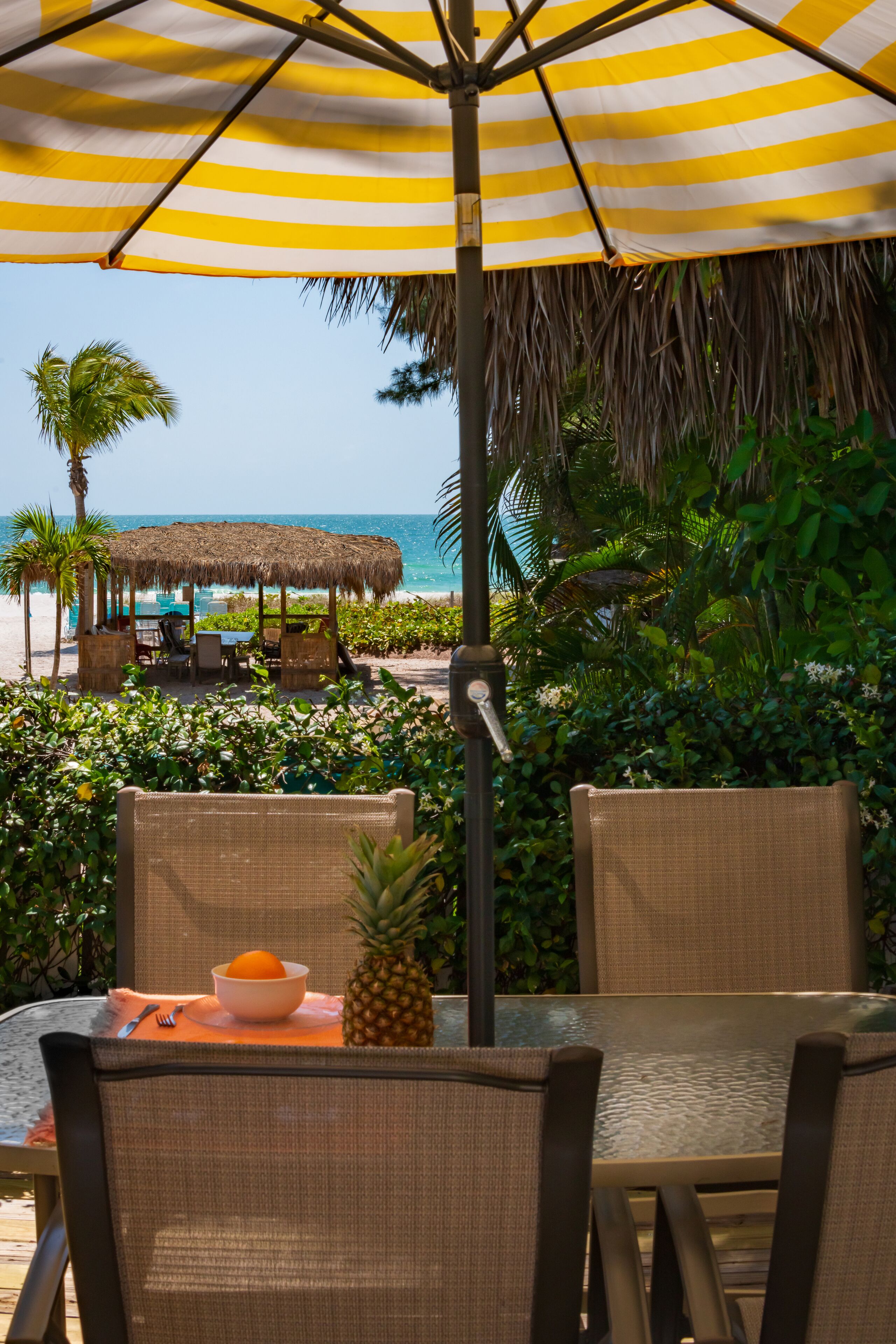 A beachside dining area with wicker chairs, a glass table, a yellow-striped umbrella, and a thatched-roof structure in the background.