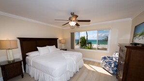 A bedroom with a large bed, a ceiling fan, a nightstand with a lamp, a dresser, and a window with a beach view.