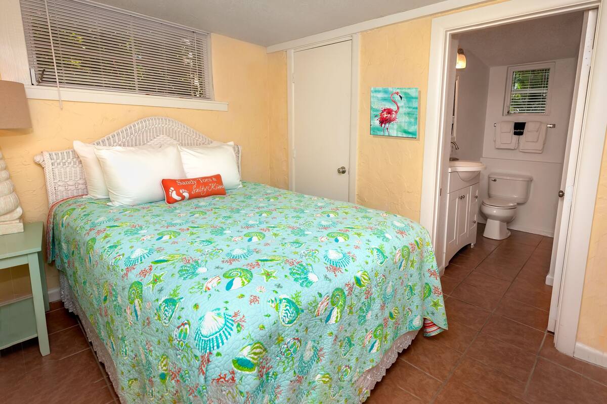 Een slaapkamer met een bed, een nachtkastje, een raam met jaloezieën, een deur naar een badkamer en een schilderij aan de muur.