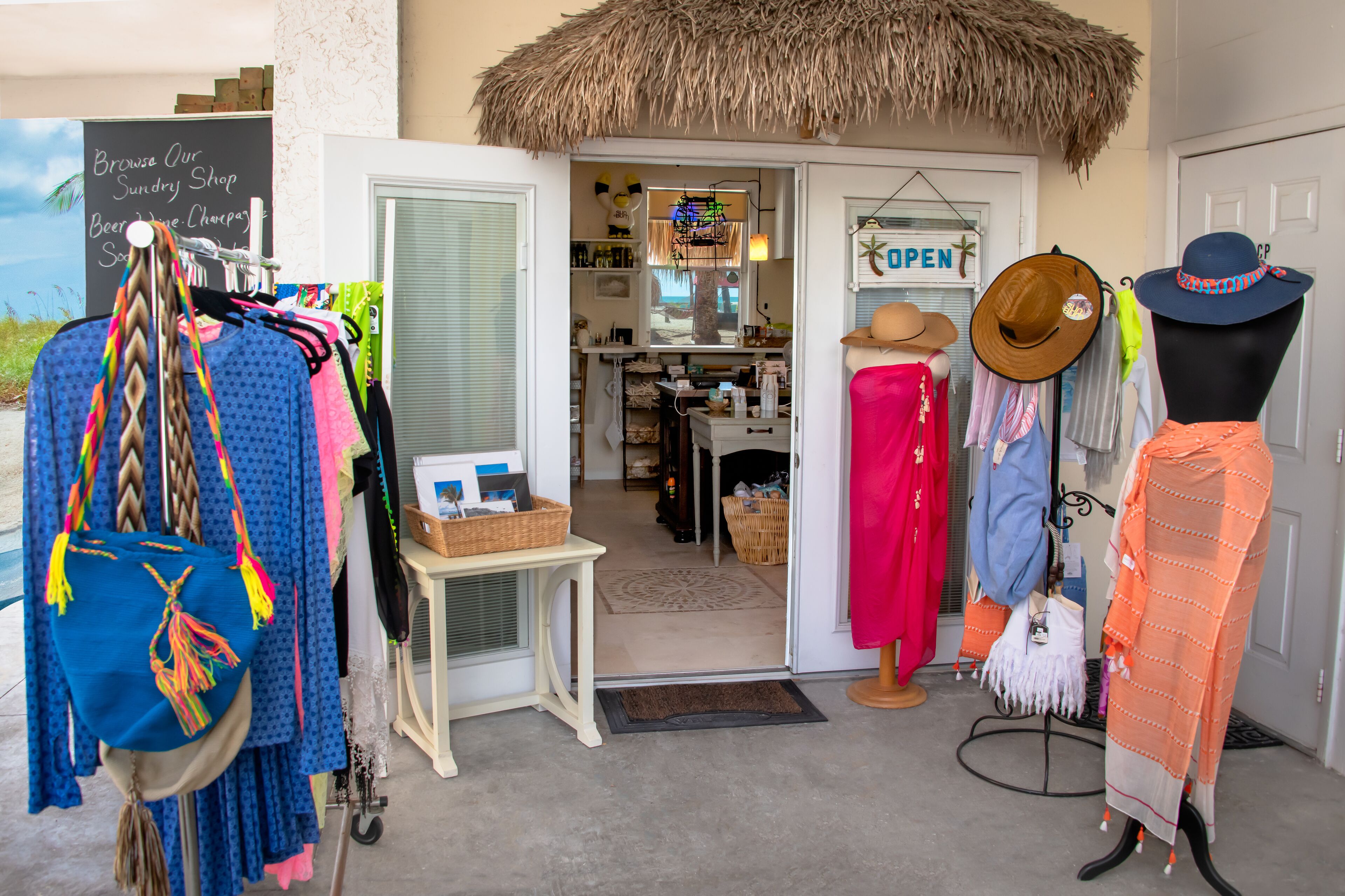 A boutique shop with a thatched roof entrance, a variety of clothing and accessories displayed outside, and a sign indicating the shop is open.