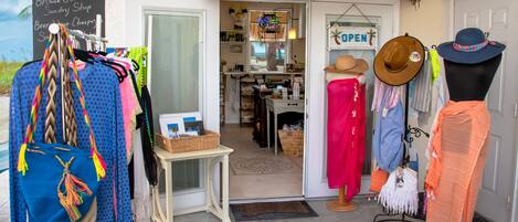 A boutique shop with a thatched roof entrance, a variety of clothing and accessories displayed outside, and a sign indicating the shop is open.