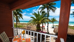 A wooden balcony with a view of a beach, palm trees, and beach chairs.