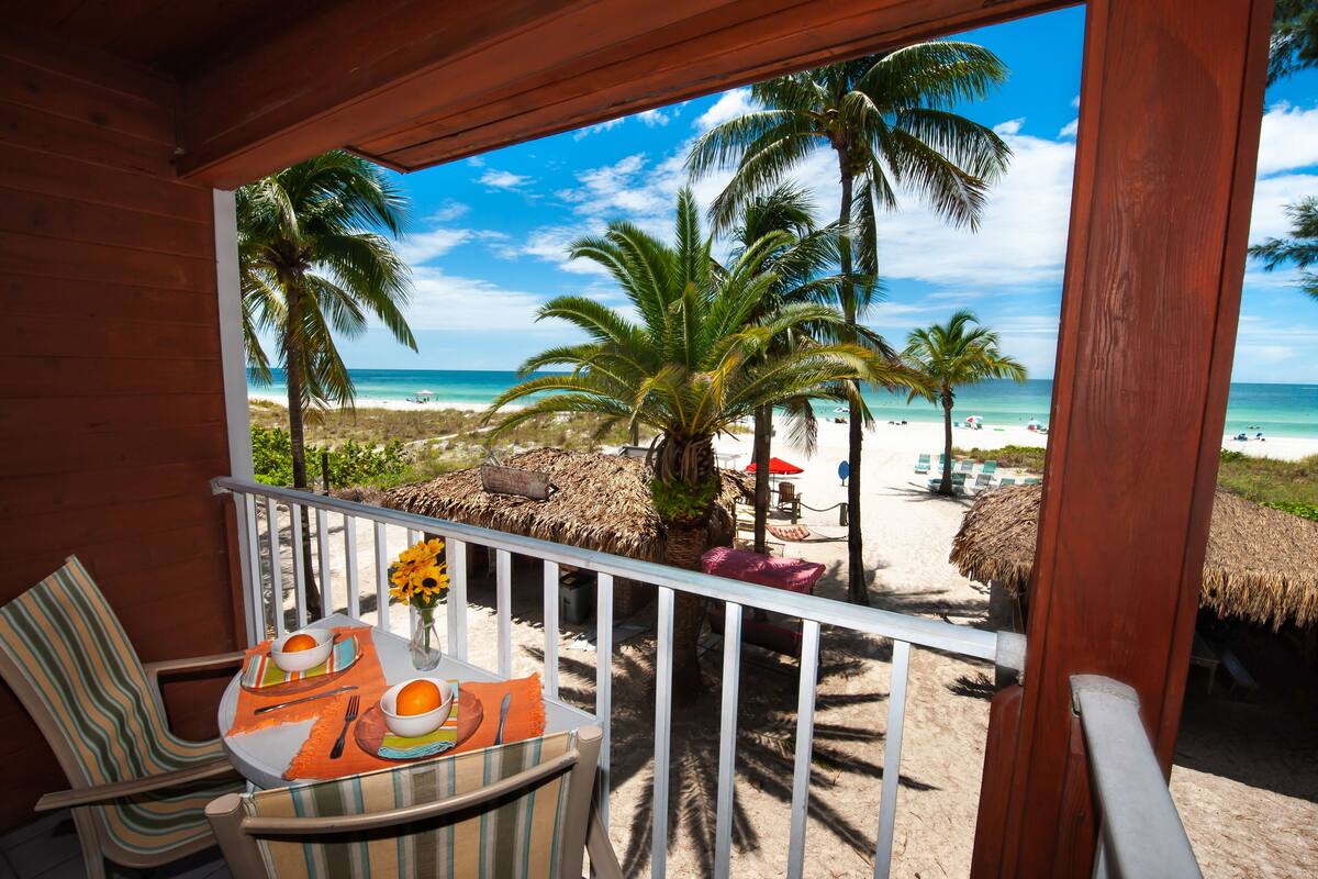 Een houten balkon met uitzicht op een strand, palmbomen en strandstoelen.