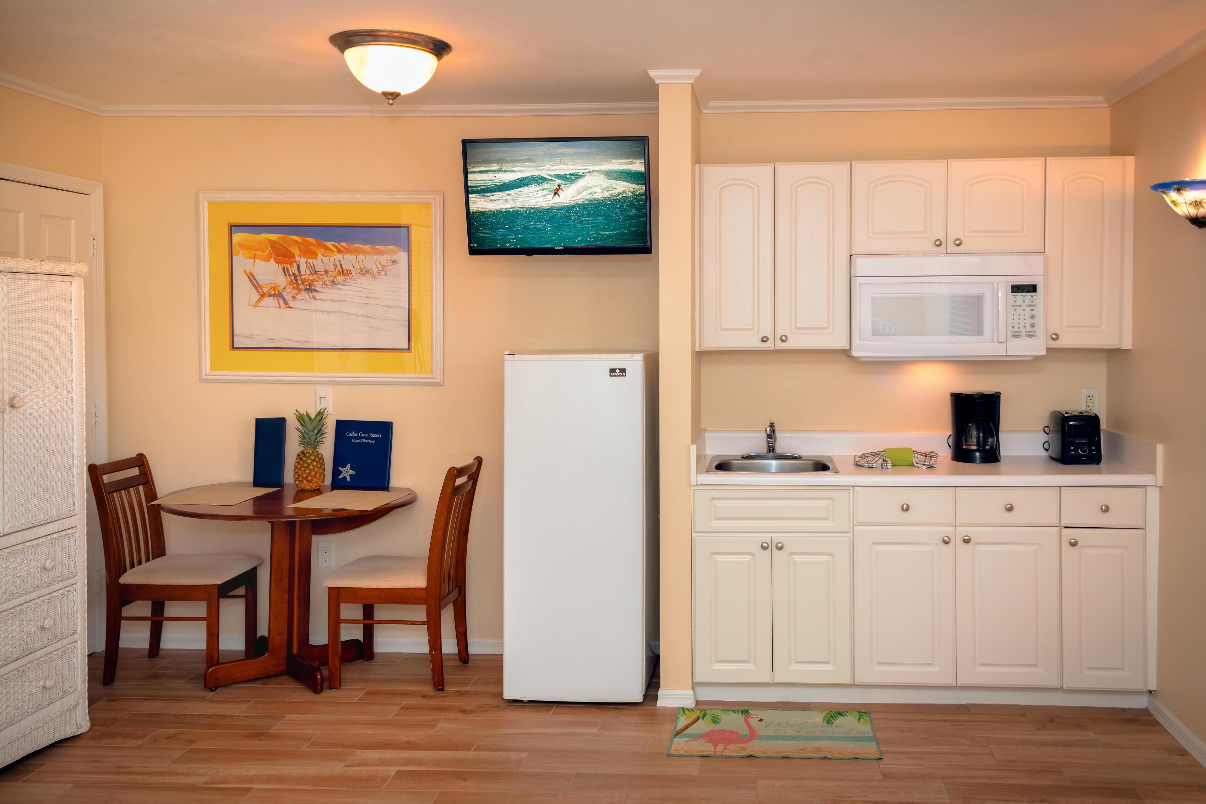 A compact kitchen with white cabinets, a microwave, a refrigerator, and a dining area with wooden chairs and a table.