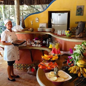 A chef standing at a buffet counter with a variety of fruits and a refrigerator in the background.