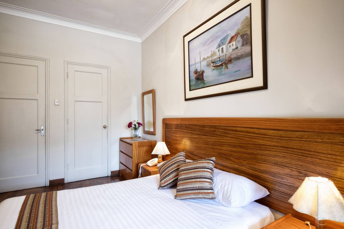 A neatly made bed with striped pillows, a wooden headboard, a bedside lamp, and a framed painting of a coastal scene.