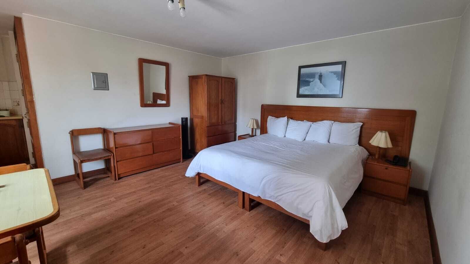 A bedroom with a wooden bed, a wooden dresser, a chair, a nightstand with a lamp, and a framed picture on the wall.