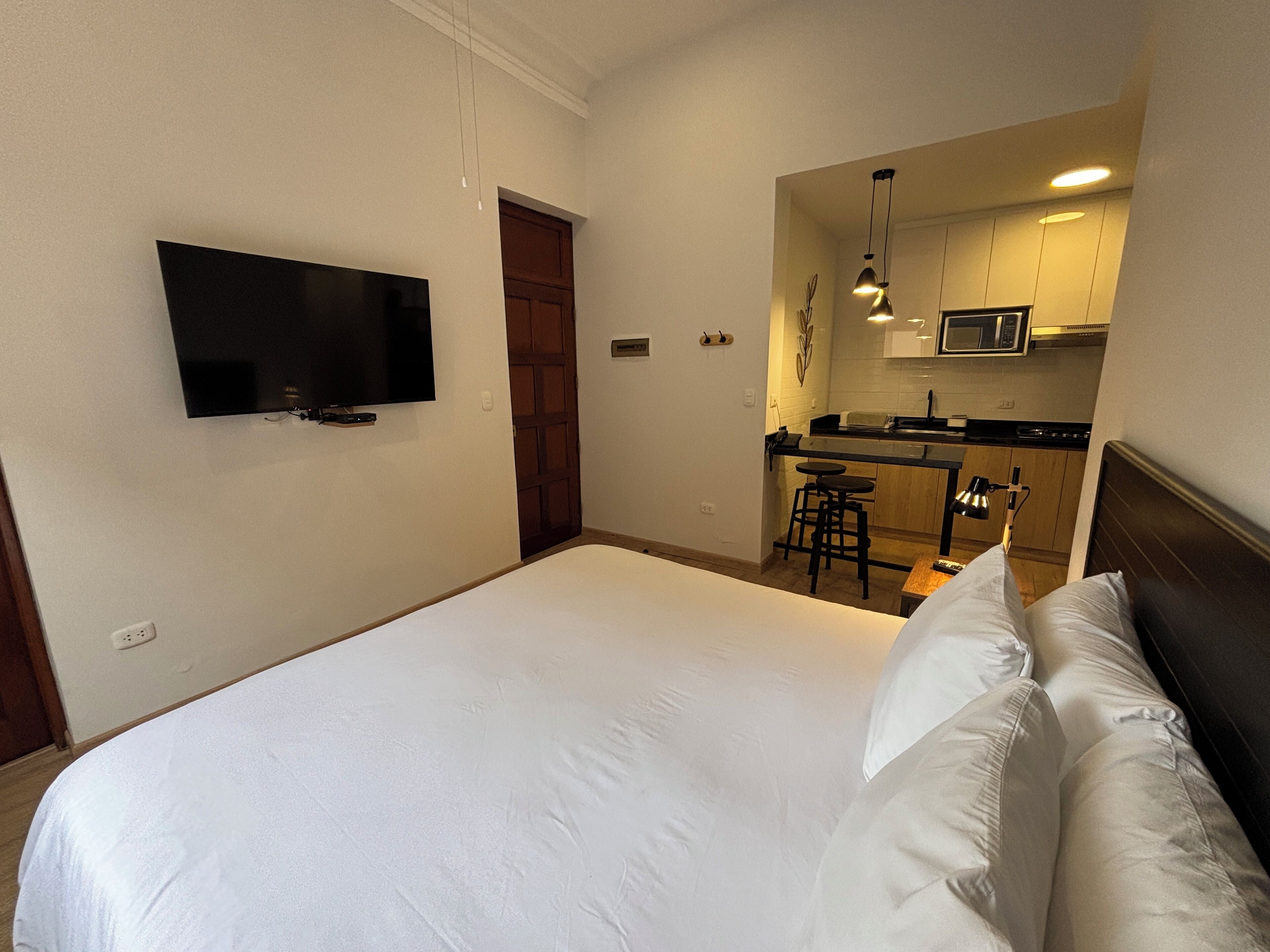 Habitación de hotel con cama, televisor montado en la pared, kitchenette con microondas y una pequeña zona de bar.