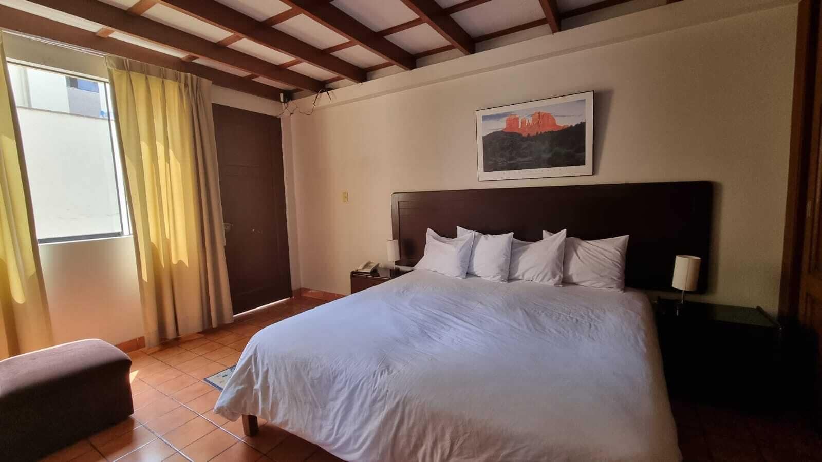 A hotel room with a large bed, a nightstand, a bedside lamp, a window with curtains, and a painting on the wall.