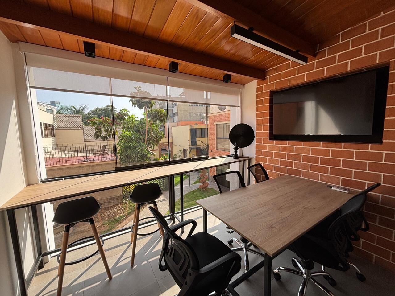 Sala de conferências com uma mesa grande, quatro cadeiras, uma tela de TV e vista através de janelas amplas.