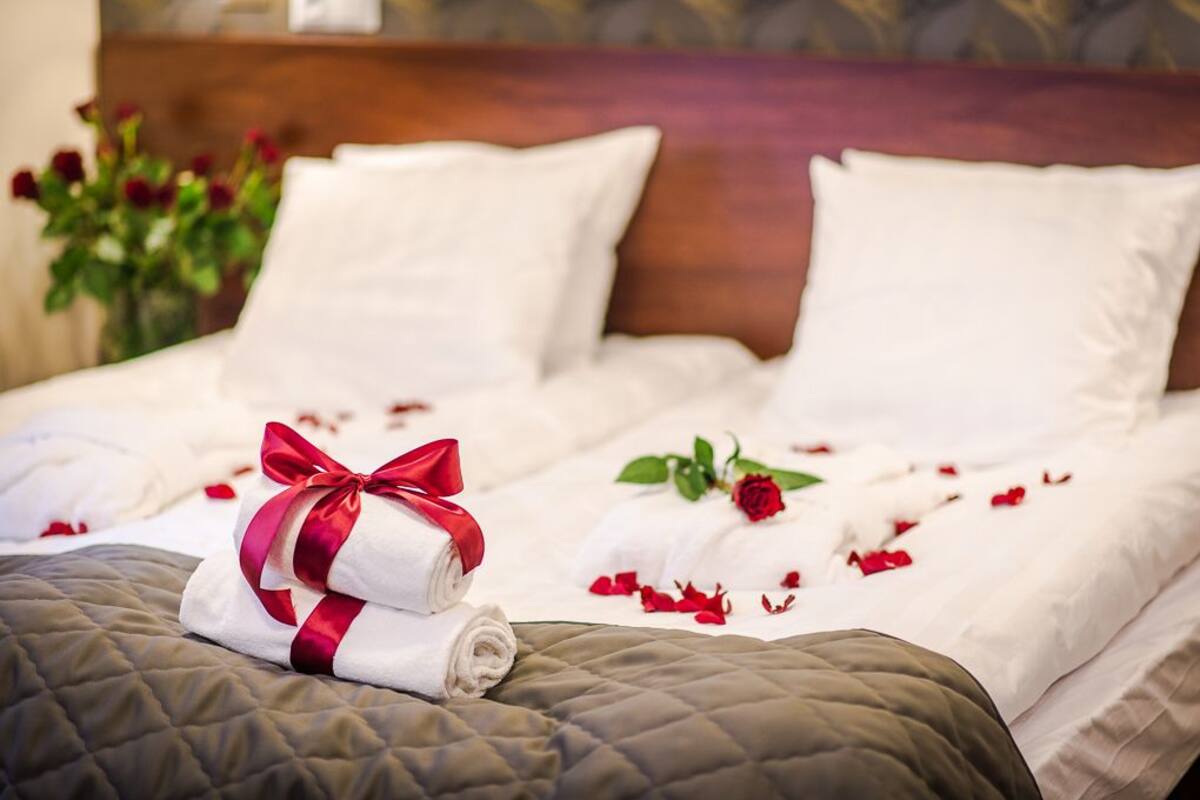 Ein ordentlich bezogenes Bett mit weißen Leinen, einem roten Band und verstreuten Rosenblüten.