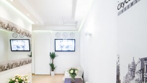 A reception area with a white reception desk, two potted plants, a sofa, and a wall-mounted TV. - City Center Rooms (Lodz)
