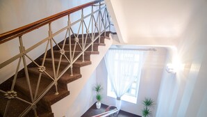 A staircase with a wooden handrail and metal balusters, leading to a landing with two potted plants and a window with sheer curtains. - City Center Rooms (Lodz)