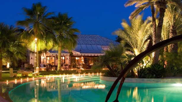 A pool area with a curved edge, palm trees, and a building with warm lighting in the background. - Lodge K Hôtel & Spa (Marrakech)