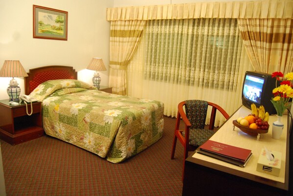 A hotel room with a bed, a desk with a computer, a chair, a telephone, a television, and a window with curtains. - Rigs Inn (Dhaka)
