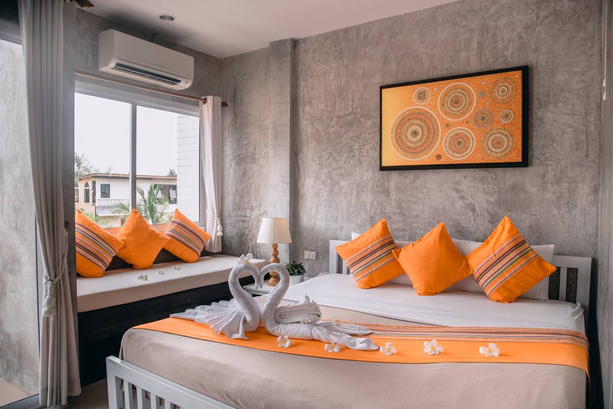 Une chambre d’hôtel avec un lit, des oreillers orange, une décoration en forme de cygne, une œuvre d’art murale et une fenêtre avec des rideaux.