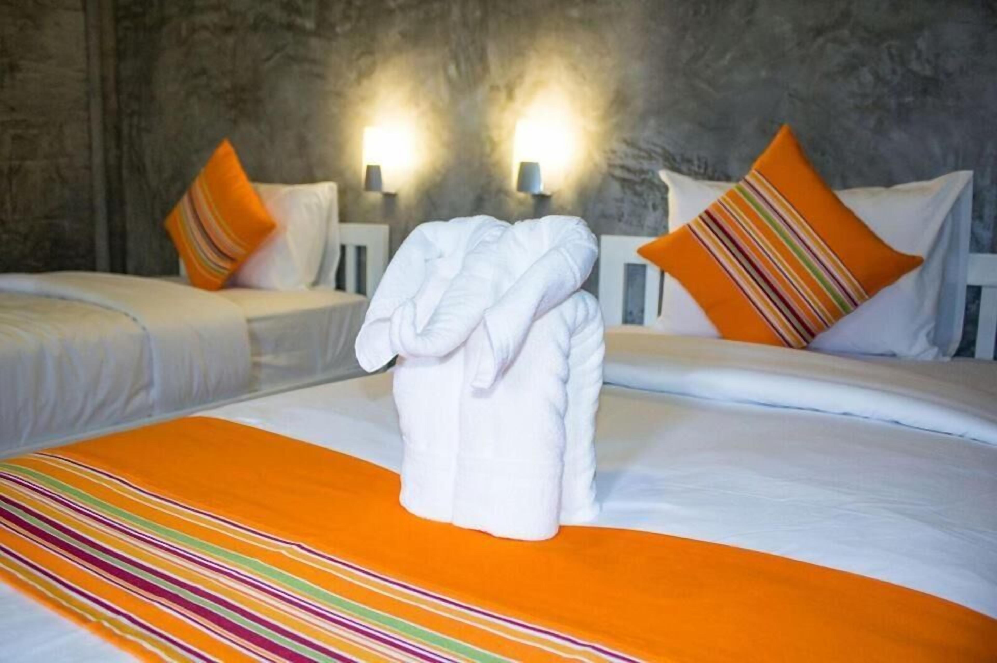 A hotel room with two beds, each with white bedding and orange pillows. There are two wall-mounted lamps providing light.