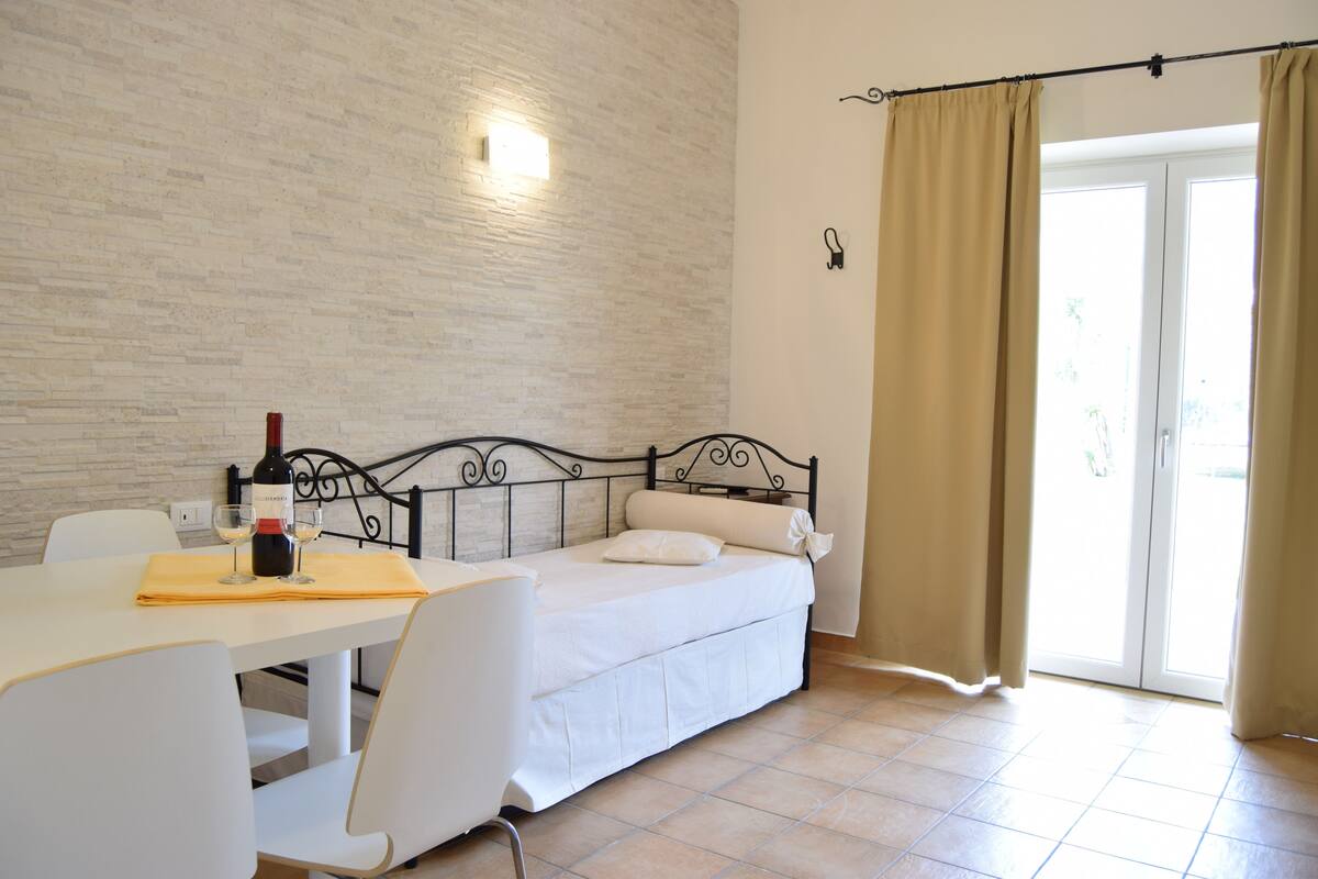 Une pièce avec un lit, une table sur laquelle se trouvent une bouteille et des verres, une chaise et une fenêtre avec des rideaux.