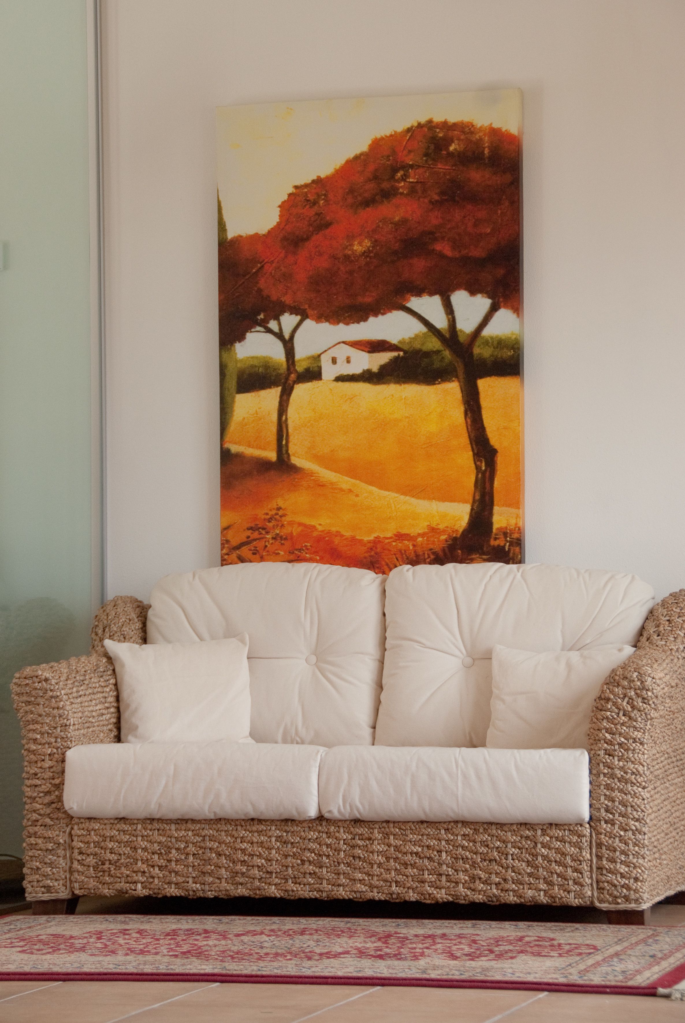 A wicker sofa with white cushions and a patterned rug in a room with a painting of trees and a house on the wall.