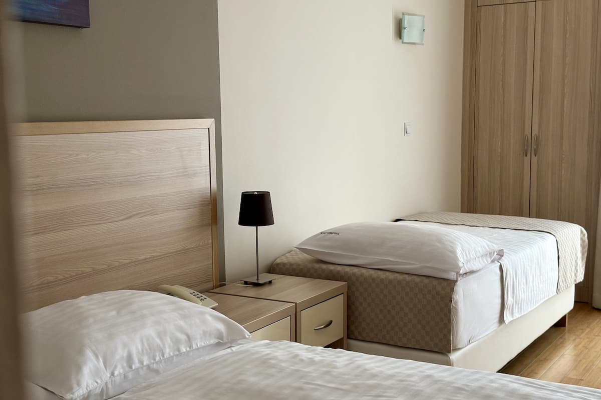 A hotel room with two beds, a bedside table with a lamp, a wardrobe, and a wall-mounted light fixture.
