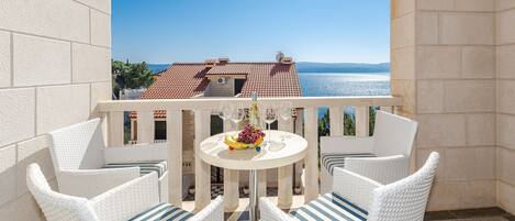 A balcony with white wicker furniture, a round table with a fruit bowl and wine glasses, and a view of a building with a tiled roof and water in the background.